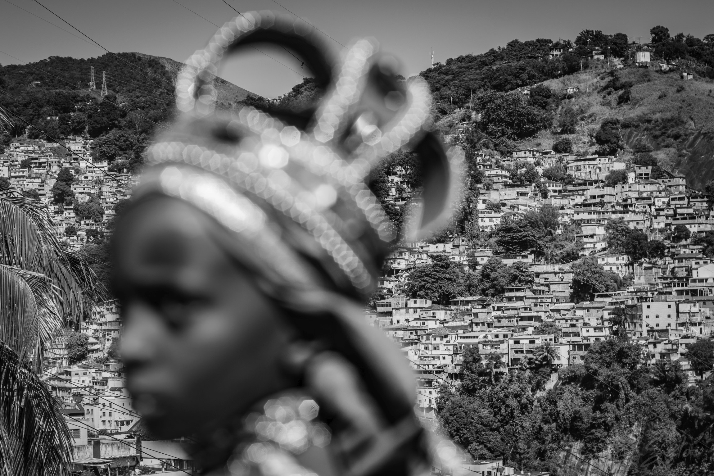 Foto em preto e branco de uma mulher negra usando uma coroa, retratada em primeiro plano desfocado e o plano de fundo em foco apresentando uma favela construída em um morro. Fotografia de Leonardo Salo.