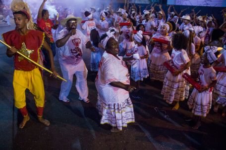 Mulheres e crianças negras de uniforme branco e saia branca tocando um instrumento de percussão vermelho em uma rua. No centro, em destaque, uma mulher negra sorridente dançando com as mesmas roupas brancas. À esquerda, um homem com o uniforme branco e de calça comprida branca cantando algo no microfone.
