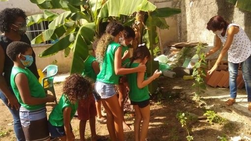 Crianças em uma horta em meio a plantas e bananeiras, com uma senhora acompanhando-as e outra senhora apontando para uma planta e explicando algo.