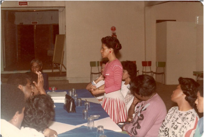 Mulher negra de camisa de manga longa vermelha em pé falando em torno de uma mesa. Na mesa sentado, homens e mulheres negras em um auditório fechado.