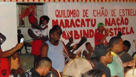 Homens negros tocando instrumentos. Ao fundo, uma mulher negra segurando uma criança olhando o desfile. Ao lado, uma parede branca com os dizeres em vermelho: "Quilombo de Chão de Estrelas, Maracatu Nação Cambinda Estrela".