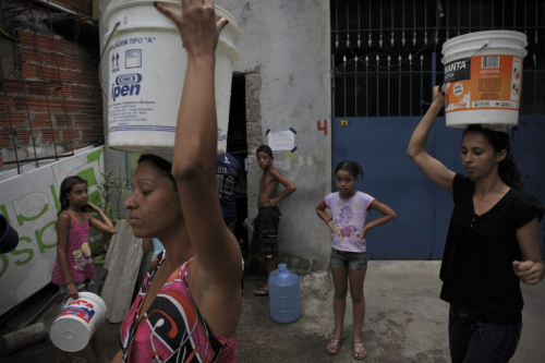 Mulheres carregam baldes na cabeça na favela de Paraisópolis, em São Paulo. Imagem: Maurício Lima/AFP