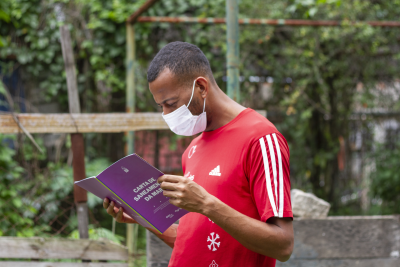 Homem de camisa vermelha e mascara facial segurando o caderno roxo da carta de saneamento da Maré